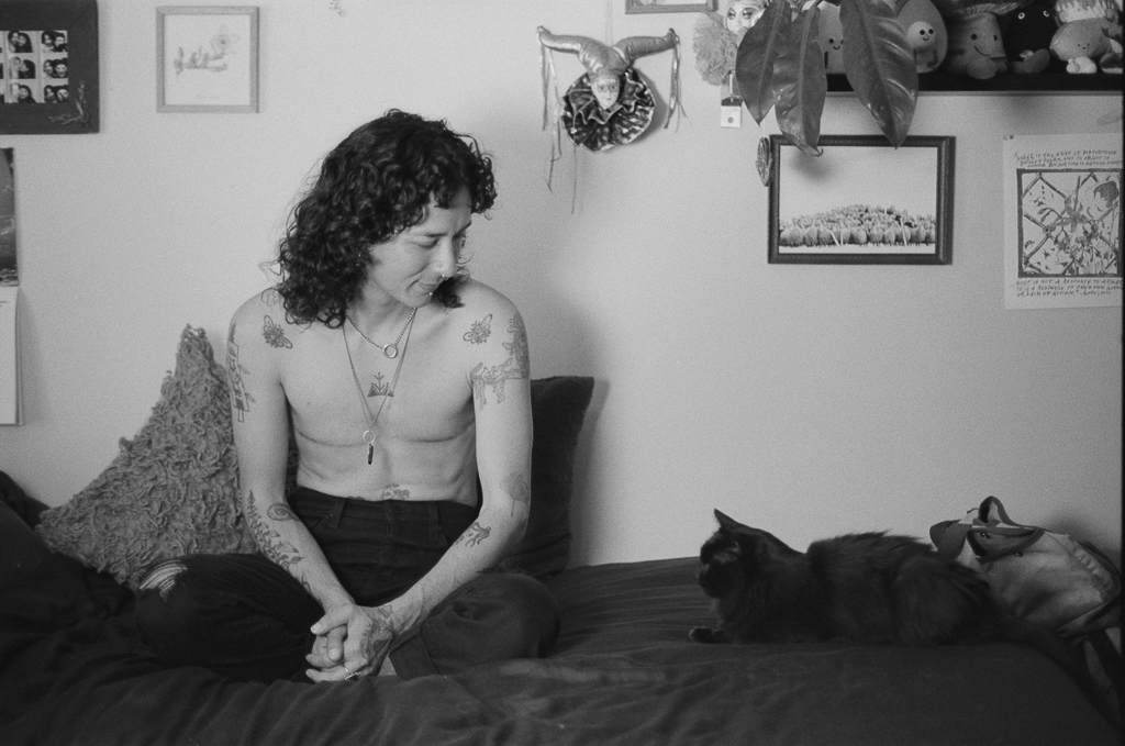 A person with long, curly, black hair sits shirtless on a couch, looking to the side at their tiny black cat lying next to them, The person has tattoos, a nippleless chest with top surgery scars, and is wearing black pants. Behind them are a fuzzy throwpillow, and various framed and textured art pieces along the wall. Their hands are clasped together in their lap.