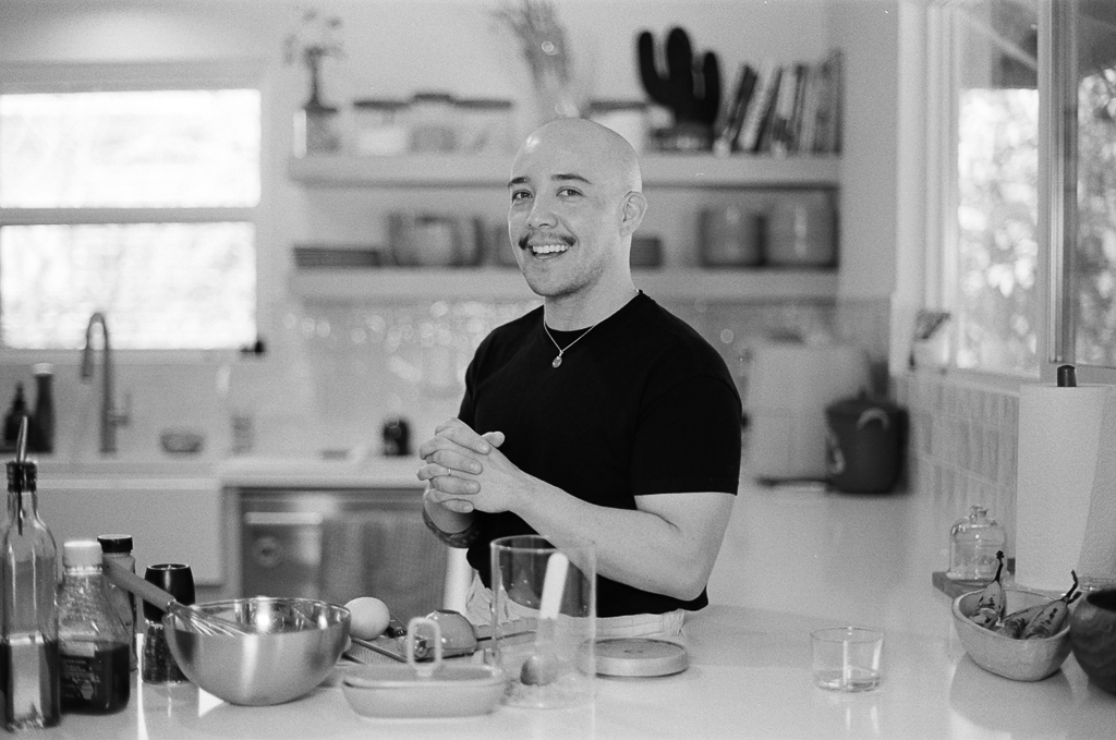 A bald, light-skinned, native Hawaiian person clasps their hands in front of their body. They have a short moustache and stand behind a bar counter, with garnishing tools and glassware in front of and behind. They're wearing a black t-shirt and smiling.