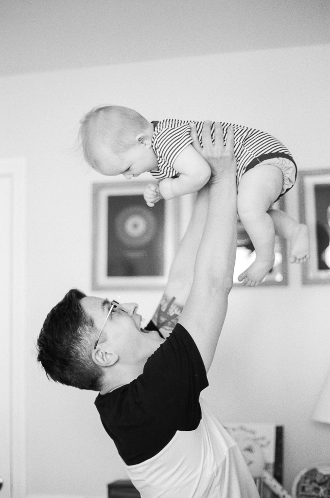 A white person with short brown hair and glasses holds their infant baby above their head, both smiling with glee. In the background, a row of paintings decorates a white wall.