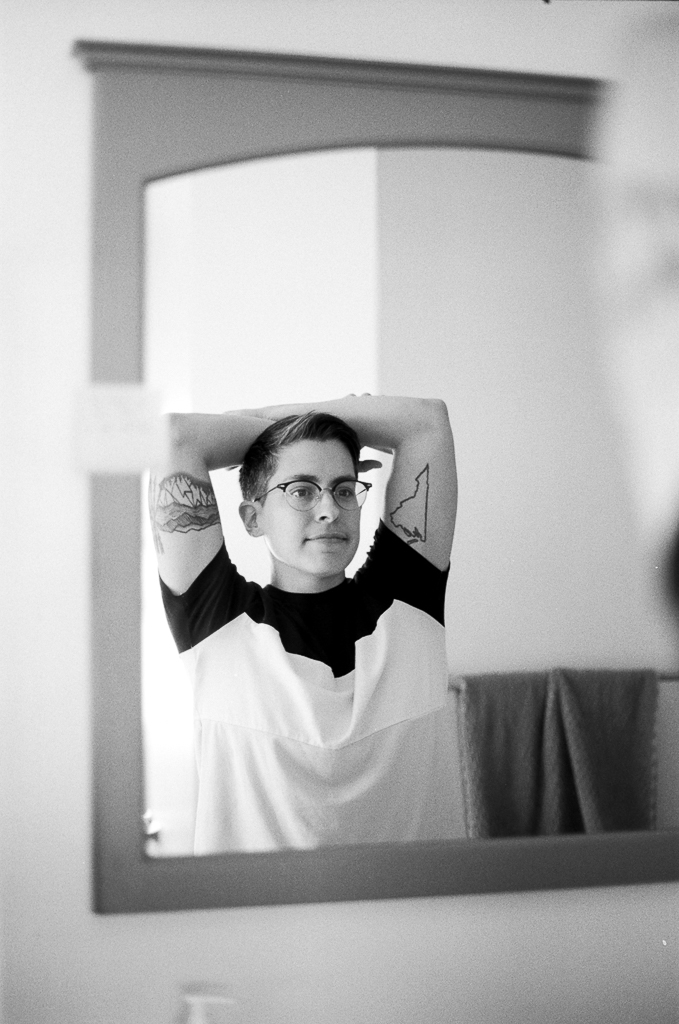 A white person with their arms lifted over their head, each hand holding the opposite forearm above their head. They're looking in a mirror, wearing a white t-shirt with black sleeves and collar, and glasses. A tattoo is visible under each upper arm.