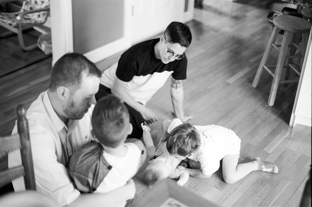 Five people—two adults, a six year old, a two year old, and an infant—spending time together on the floor of a house. The adult on the left has short hair and a button-down shirt, long sleeves rolled up past the elbow; they hold the six-year-old in a hug. The other adult watches over the 2-year old child giggling at the infant. The group seems calm and focused on one another.