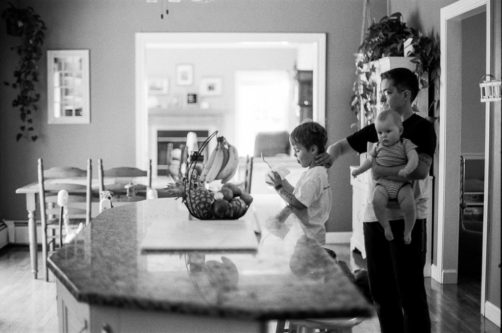 A white person with short hair holds an infant in their arms, their hand resting on the back of the neck of a small child in front of them. They're at a standalone kitchen island in a well-lit kitchen, beautiful artwork and plants adorning the space around them.