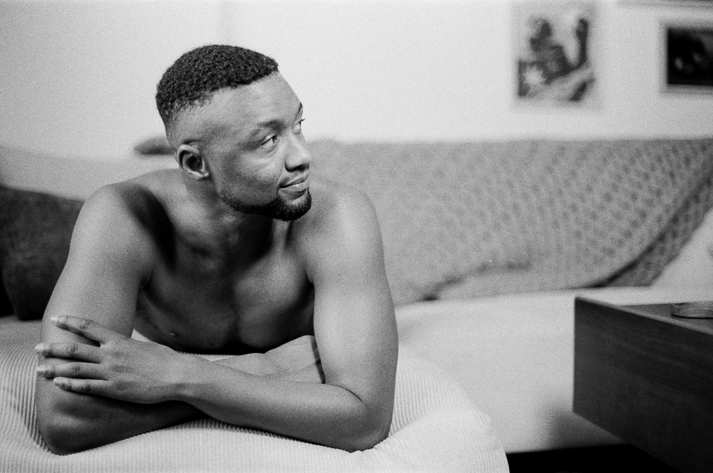 A Black trans man, shirtless, positioned face-down on the chaise of a couch, his back arched slightly upward and arms crossed. He looks off to the side. He has facial hair just along his chin, and short black hair buzzed in a fade.