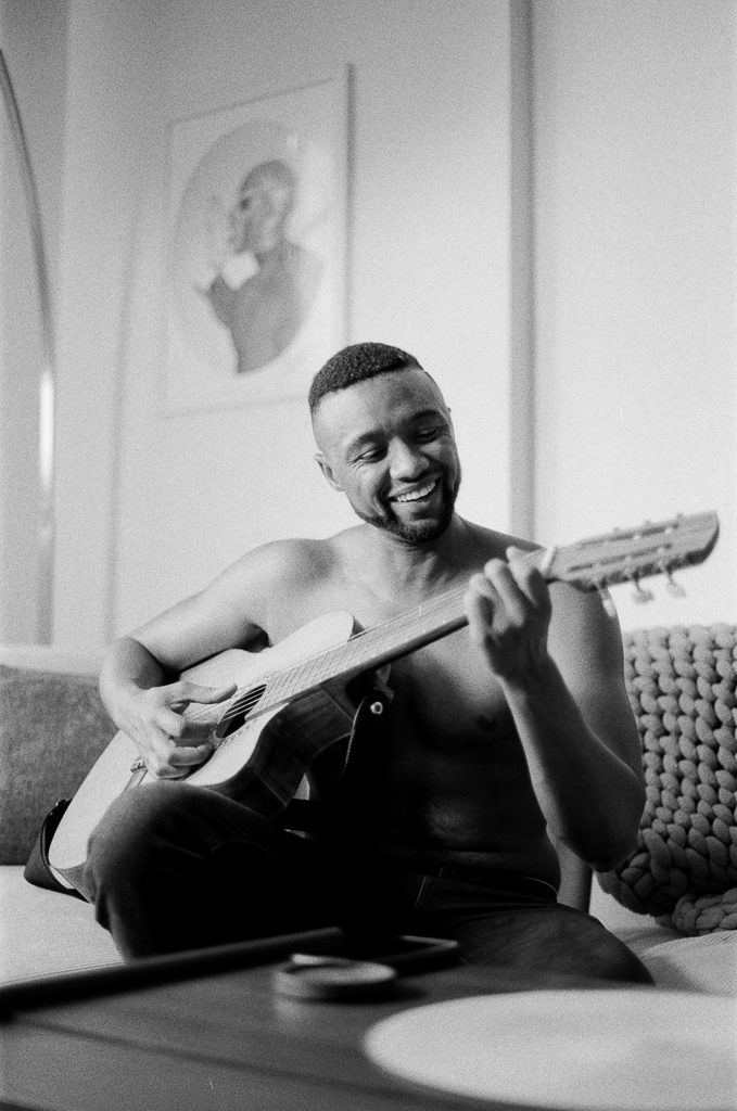 A Black trans masculine person, shirtless, smiles while playing a guitar and sitting on a couch. A crocheted blanket made with thick yarn sits behind him, and a coffee table appears in the foreground.